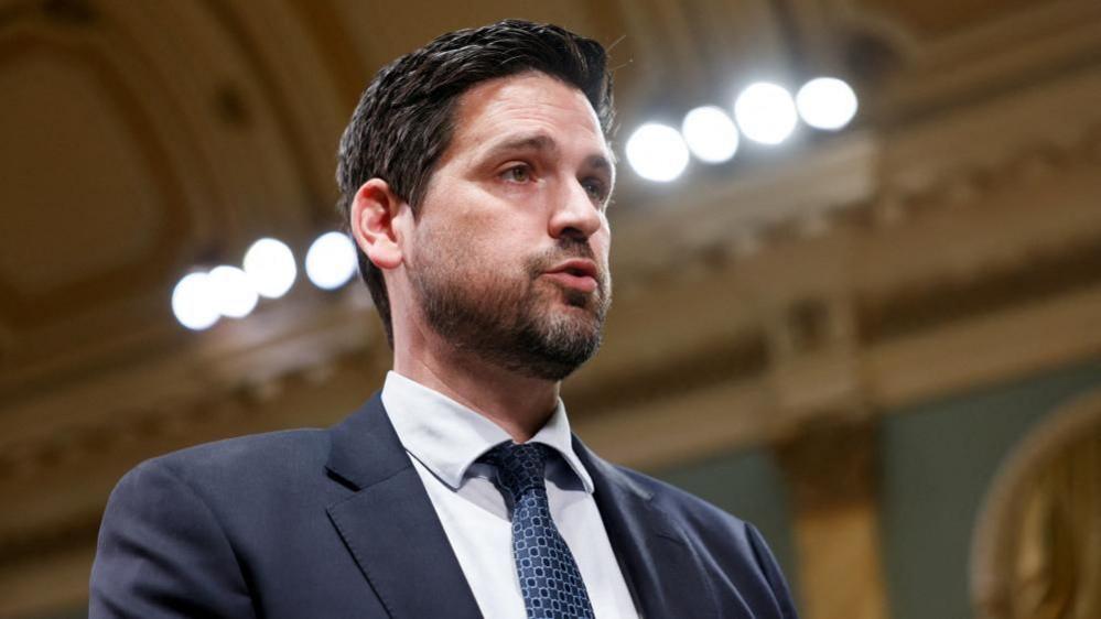 Sean Fraser, with short cropped dark hair and a beard, seen in a two-thirds profile. He is speaking and wears a navy suit and patterned blue tie. 