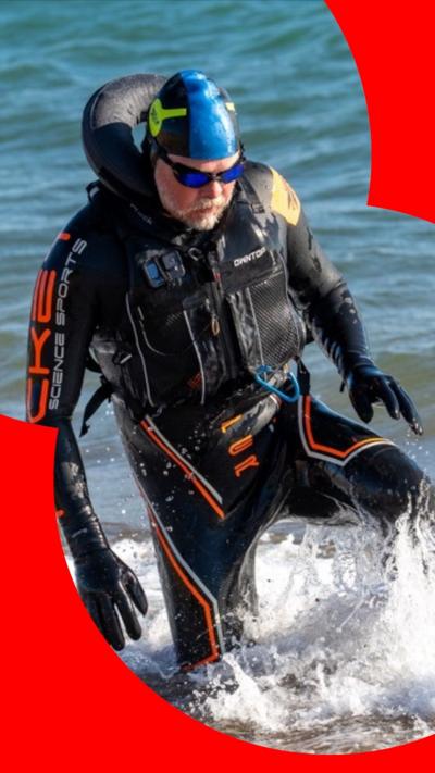 A man with a white beard and moustache wading out of the sea in a wetsuit. He is a wearing goggles and a blue head covering.