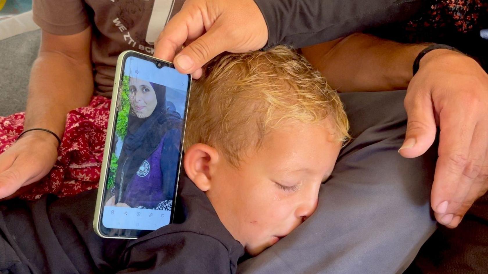 Hands hold up a mobile phone showing an image of Fida al-Toutah, wearing a black hijab, as a young boy next to the phone turns his face away in distress
