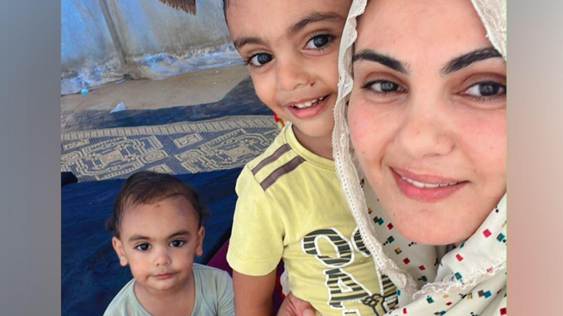 Amal Alabadla sits wearing a spotted headscarf, with her arm around one son in a yellow t-shirt and her younger son sat in the background holding a flask. All of them are sat inside a tent in Gaza