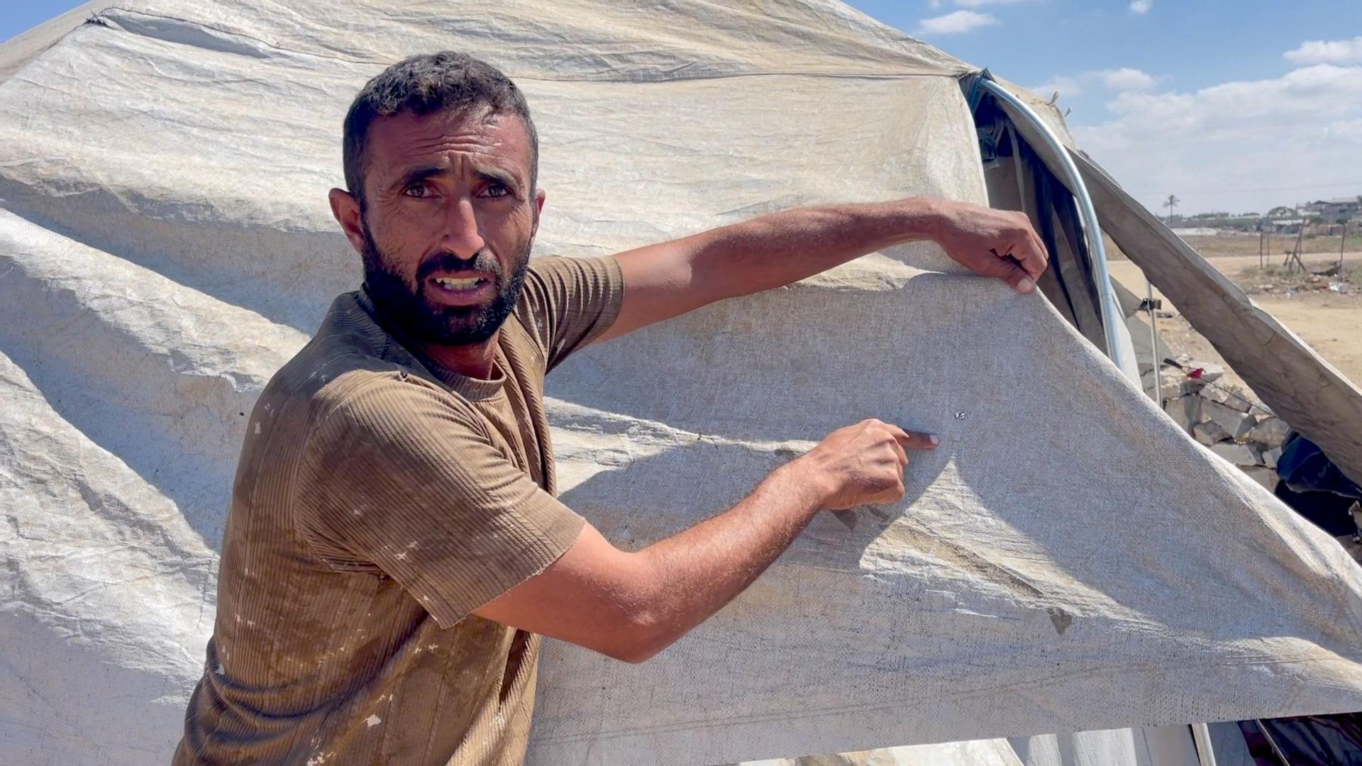 A bearded man in a beige t-shirt points a piece of tent cloth he's holding up, in the Gaza Strip