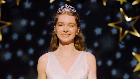 A still from Carrie shows the main character smiling as she stands on a stage wearing a tiara and a white prom dress. Gold and white stars fill the background behind her, and a spotlight illuminates her.
