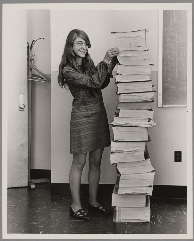 A black and white photograph of a woman standing next to a pile of books that is as tall of her.