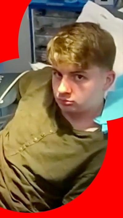 A young man with blond hair sits up in a hospital bed. He is wearing a long-sleeved, olive-green shirt.
