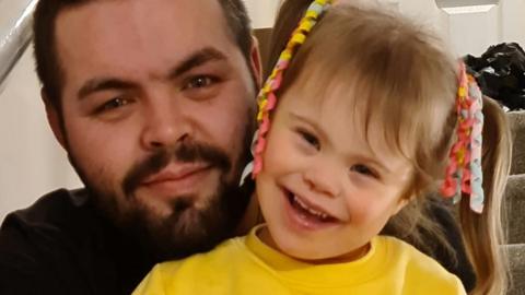 Aaron Huxter (left) with his daughter Olivia-Grace. Aaron has blue eyes, short brown hair and a bushy beard. Olivia wears a yellow jumper, has her brown hair in pigtails and smiles at the camera. 