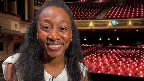 Beverley Knight is wearing a white top and a clip-on microphone sits on the stage at a theatre, with rows of red auditorium seats visible in the background.