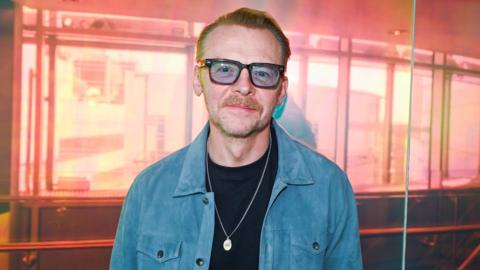 Simon pegg in a blue shirt and blue tinted glasses in front of a pink background
