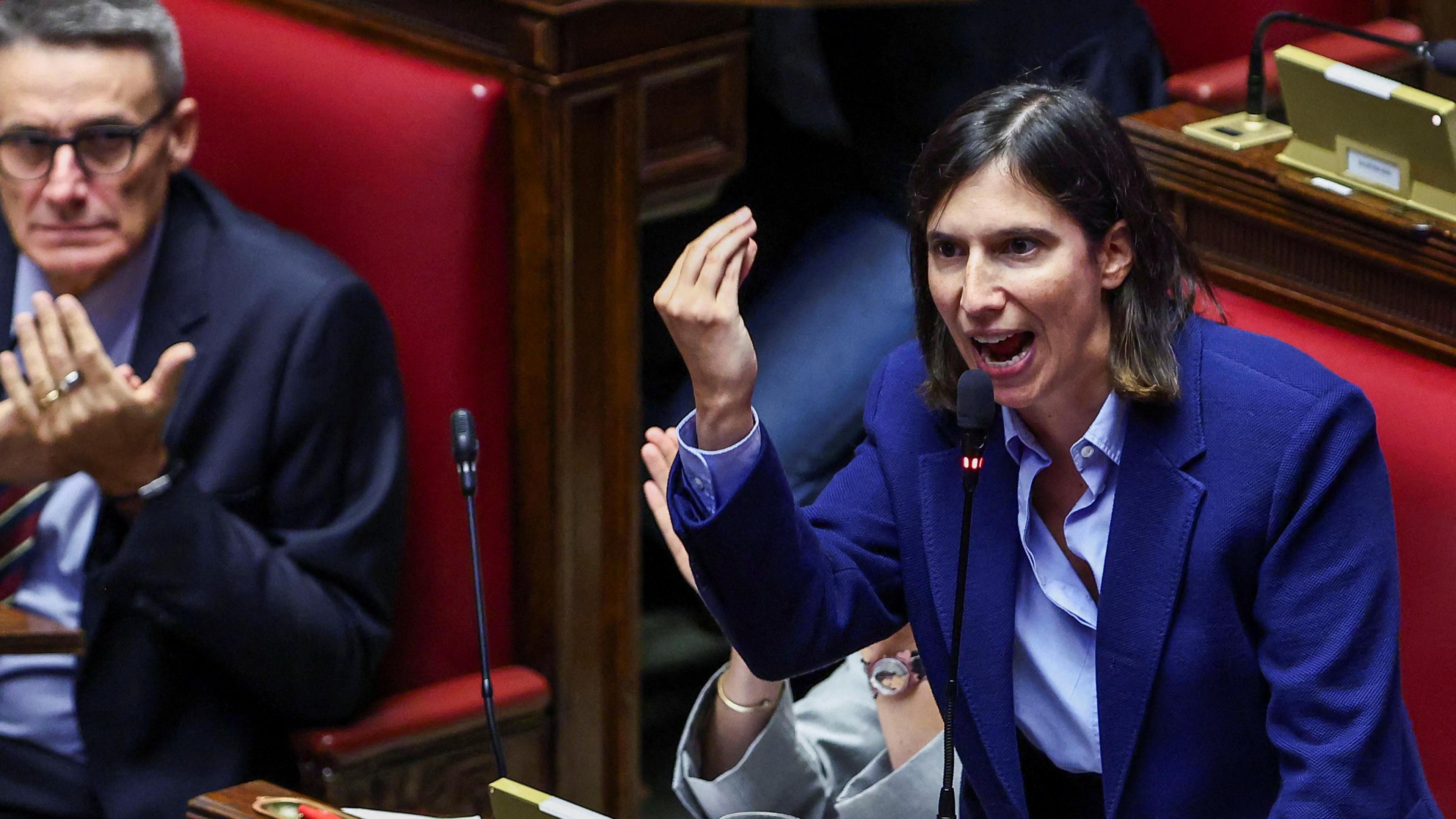 Elly Schlein speaks in parliament. She wears a dark blue blazer above a light blue buttoned shirt. She has short brown hair. She is speaking into a black microphone and gesticulating with her right hand.