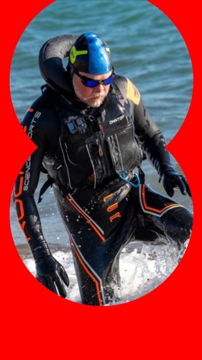 A man with a white beard and moustache wading out of the sea in a wetsuit. He is a wearing goggles and a blue head covering.
