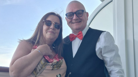Paul Scanlon and his wife Deborah standing on the balcony of a cruise ship. They are a middle-aged couple - Paul is bald and is wearing dark, circular glasses, a white shirt, black waistcoat and a red bow tie. He has his arm around Deborah who has horn-rimmed Ray-Ban sunglasses on and has dark shoulder-length hair. She is wearing a peach, sleeveless dress with a leaf pattern on. 
