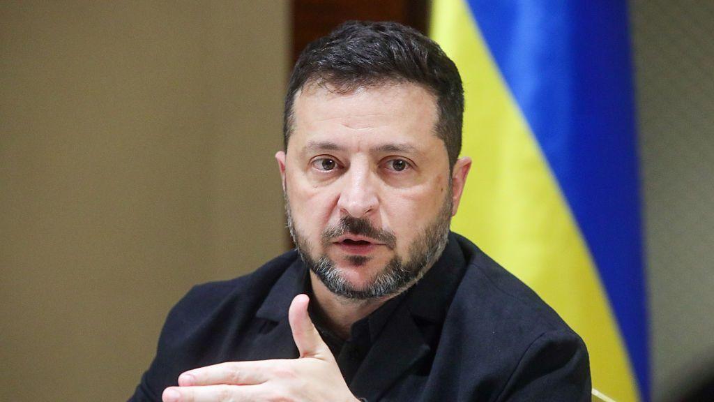 Ukrainian President Volodymyr Zelensky speaks to journalists. He is gesturing and sitting in front of the Ukrainian flag.