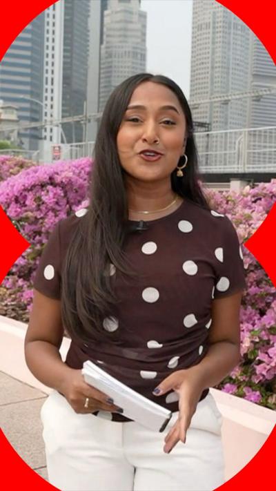 A reporter with long brown hair stands in front of a flower bed in Singapore. She is wearing white trousers, and a brown T-shirt with white polka-dots.