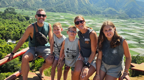 Family of five sitting on a wooden fenced ledge with rice padis in the background and green hills behind . Tom is 38, white, short fair hair. Morgan and George have short fair hair. Dani has long fair hair in a ponytail and Olivia has loose long dark blonde hair. All five wear shorts and sleeveless tshirts in varying shades of grey. Dani and Tom wear sunglasses. They all smile at the camera.