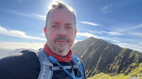 Simon Morris takes a selfie on Yr Wyddfa with the summit in the background while on a sunny day