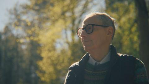 Ian Kirby in Chopwell Woods in Northumberland. He is wearing glasses, a multi-coloured jumper, a navy gillet and a blue shirt. He has short grey hair. There are tall trees behind him and the sun is shining on his head.