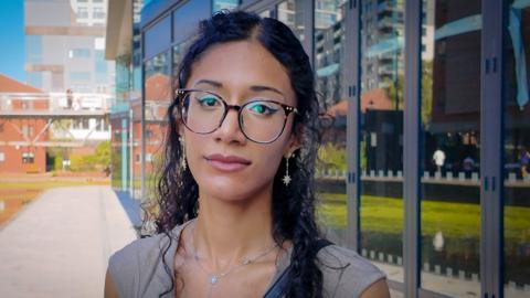 Aaliyah stands outside in front of a building. She has long wavy hair and is wearing glasses