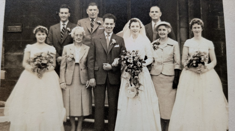 A black and white picture of a bride and groom on their wedding day with two bridesmaids and several other people.
