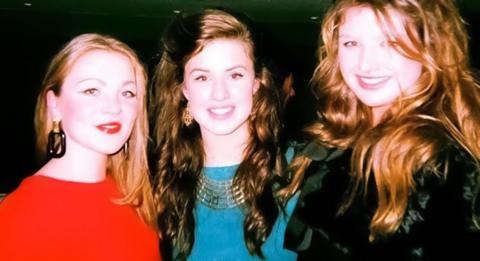 Three women standing close together. all have long brown hair. The woman on the left is wearing large earrings, red lipstick and a red top. The woman in the middle is wearing matching metal earrings and necklace and a light blue top. The woman on the right is wearing a black top with a vblack silk parter detail. They are all smiling.