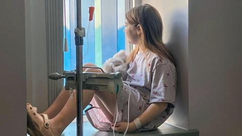 A little girl of around 13 years old with long straight brown hair tucked behind her ears sits half silhouetted in a hospital window attached to a drip with a teddy bear in her lap. She is looking away from the camera and out of the window.