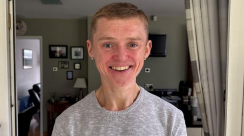 A man smiling at the camera. He is wearing a grey jumper