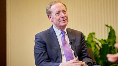Microsoft president Brad Smith sitting with his hands clasped in his lap wearing a blue suit, white shirt, and purple-patterned tie. Behind him in soft focus, there is a beige background and some green plants.