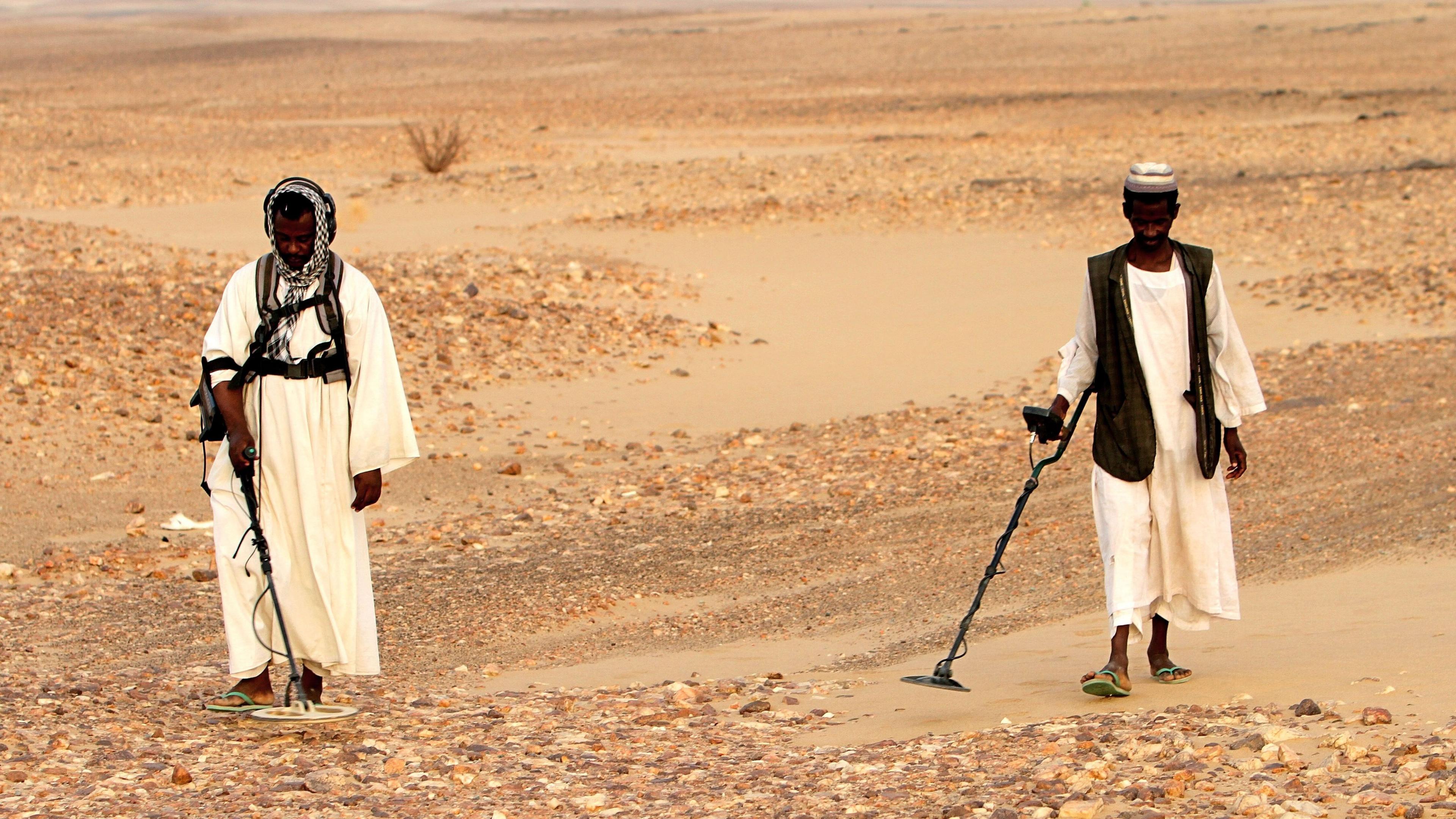 Sudanese men wearing traditional clothing search for gold with metal detectors in the desert.