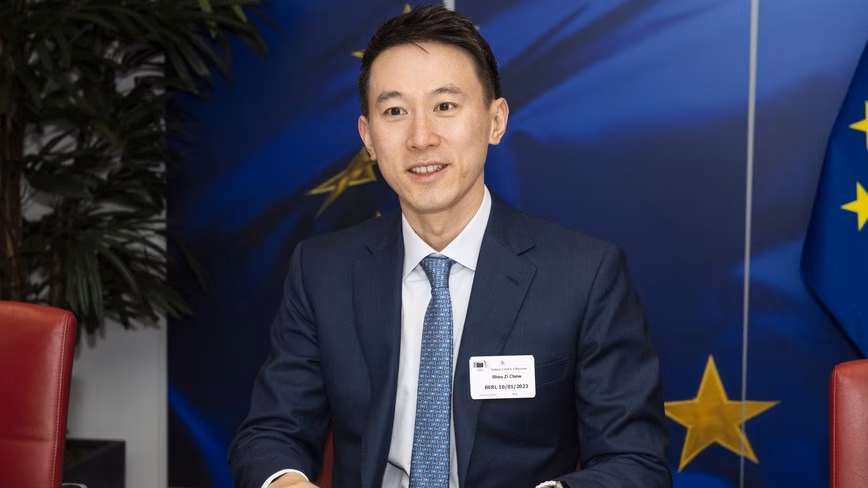 Shou Zi Chew in a navy suit and blue tie, smiling at a meeting table in front of a large EU flag backdrop