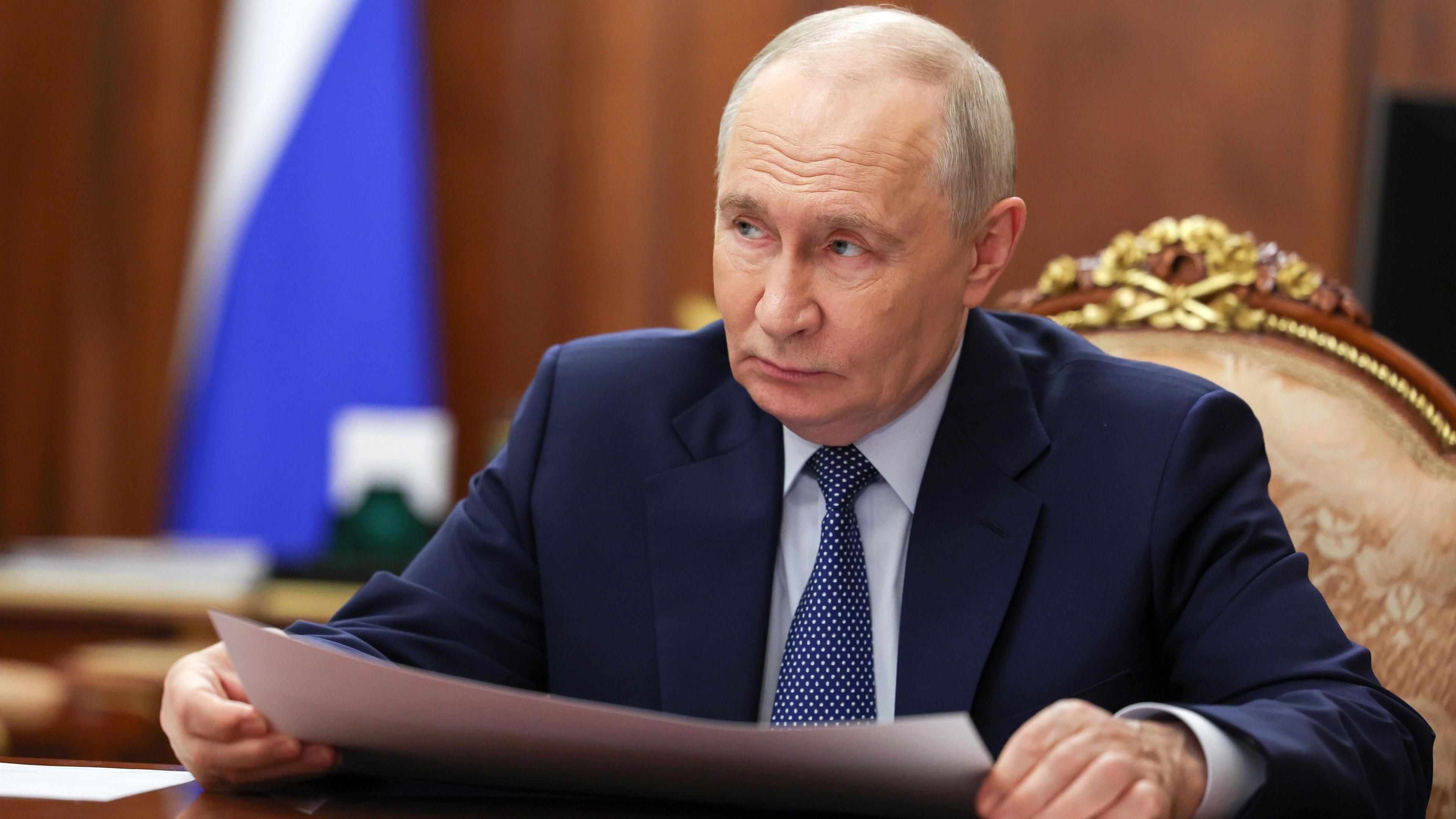 Russian President Vladimir Putin looks to the side as he sits at a wood table. He is wearing a blue suit and tie and holding a piece of paper