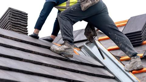 Lower half of a workman standing on a roof with a chimney visible. Some tiles are not yet laid on the roof structure.