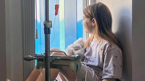 A little girl of around 13 years old with long straight brown hair tucked behind her ears sits half silhouetted in a hospital window attached to a drip with a teddy bear in her lap. She is looking away from the camera and out of the window.