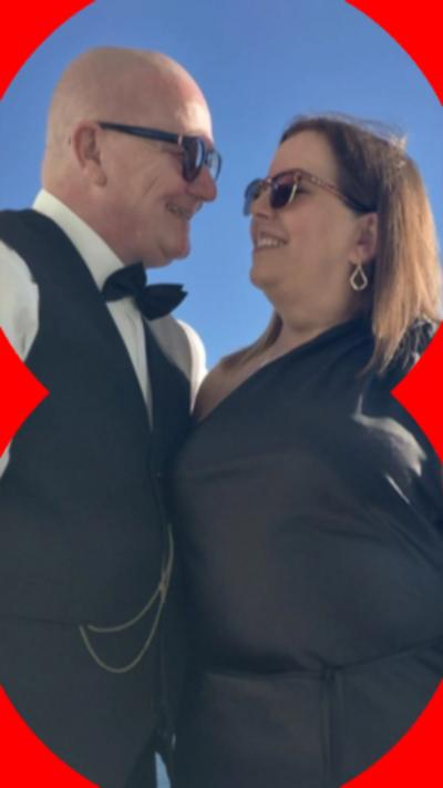 A man (left) and a woman (right) smiling and looking at each others faces,. They're both wearing sunglasses. The man has  is wearing a waistcoat and bow tie and the woman is wearing a black dress.