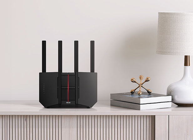 An Asus RT-BE92U router with four antennas on wooden furniture, next to a lamp and some books