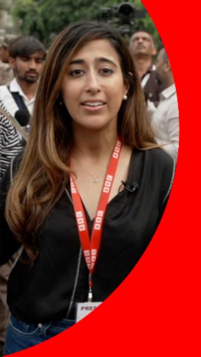 BBC Correspondent, Azadeh Moshiri, has long brown hair and is wearing a black shirt with a red lanyard 
