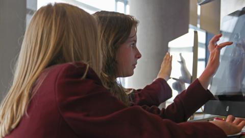 Three students use touchscreens in a close-up. Only one girl's face is visible as she stares intently at the screen.