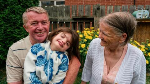 A young child is sitting on an older man’s lap, both are smiling. An older woman is smiling at them. There are yellow flowers, grass and a fence in the background. 