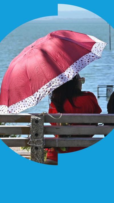 A woman sits on a concrete bench facing the sea. She is wearing red and shading herself under a red umbrella.