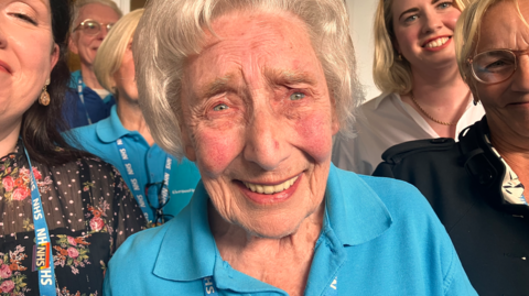 Image shows Audrey, a 96-year-old lady wearing a blue hospital volunteer t-shirt. She is surrounded by younger women wearing lanyards and smiling at the camera. 