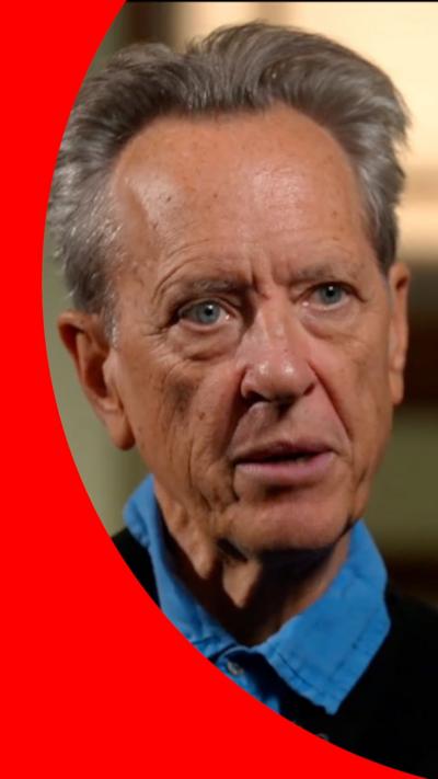 Actor Richard E Grant wearing a dark coat and light blue collared shirt