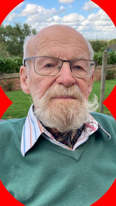 A photo of an older man with a full white beard and moustache, outside on a sunny day. He is wearing glasses, a white shirt with a blue and red striped collar, and a V-necked teal jumper.
