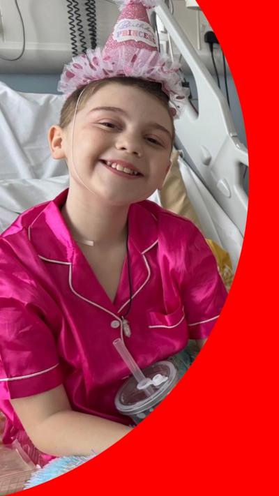 A girl sits in a hospital bed. She is wearing pink pyjamas and a pink birthday hat, and it sitting underneath a multi-coloured fluffy blanket.