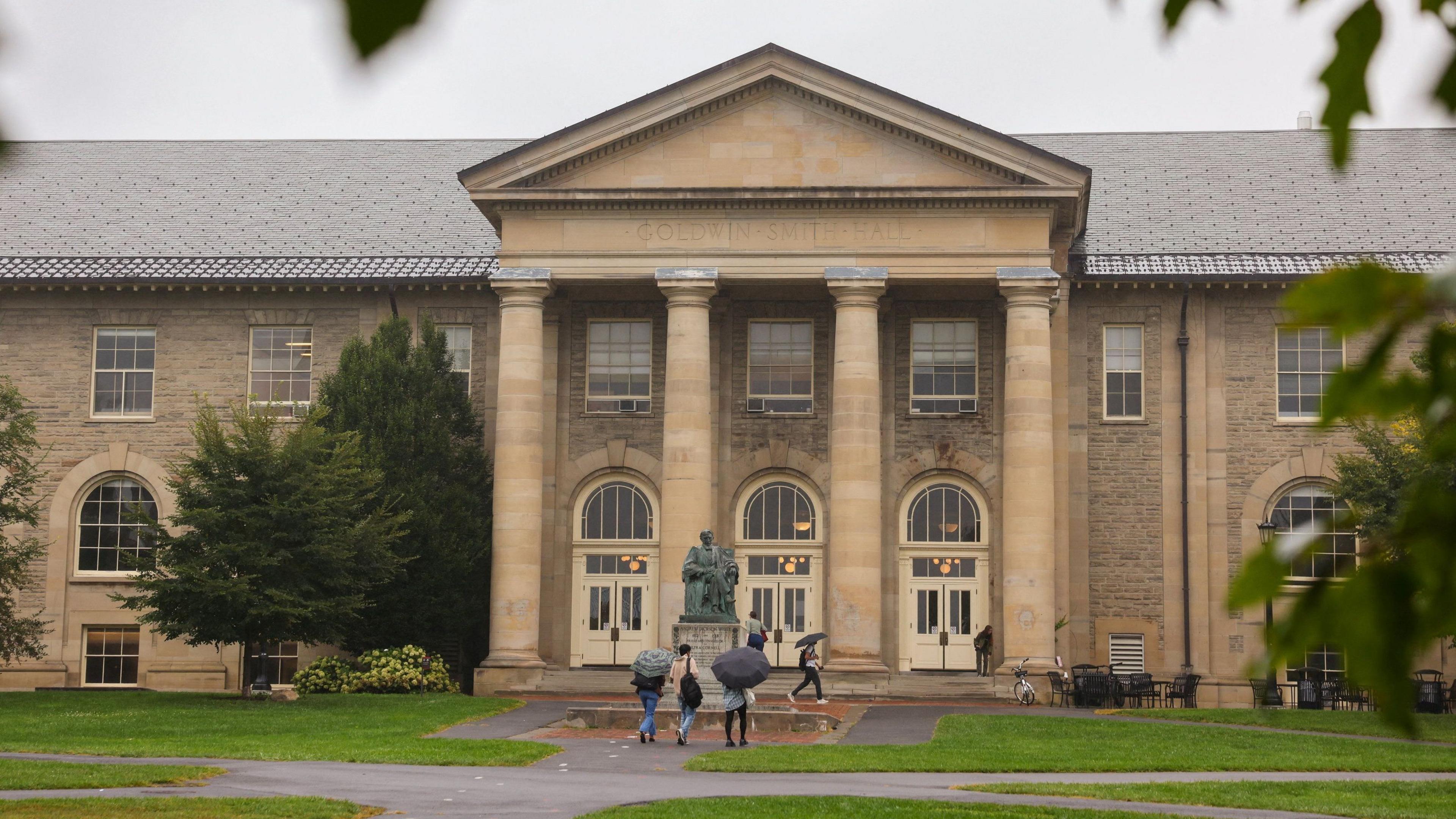 Students holding umbrellas walk towards Cornell University, built in yellow stone with four pillars at the entrance.