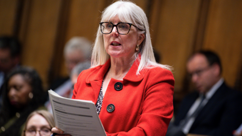 A female MP with shoulder length blonde hair and glasses standing up to deliver a speech in the House of Commons. She is wearing a red jacket and has papers and a pen in her right hand.  