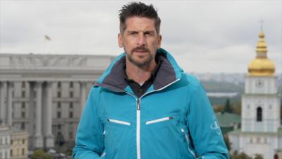 Man in a blue windbreaker standing in front of Kyiv landmarks