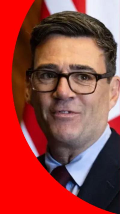 Andy Burnham has short black hair and is wearing black gasses, a dark suit with a purple tie.