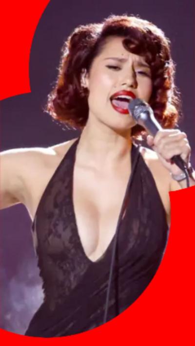 Singer Raye, has long red hair, she is wearing a black dress. She is singing on stage with a microphone in her left hand.