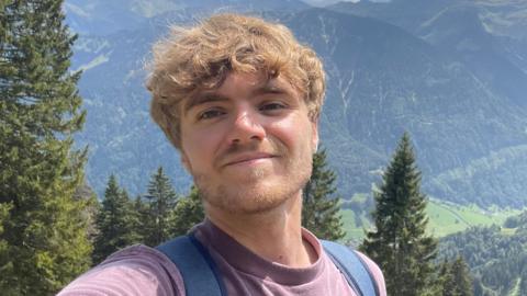 A selfie style picture of Tom standing in a lush green landscape with mountains in the background. The 24-year-old wears a purple T-shirt and backpack.  