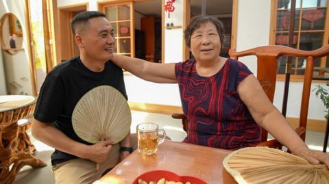 A man and elderly woman sitting at a table together, the woman's hand is on the man's shoulder and they are smiling