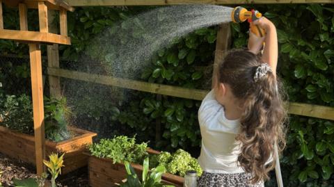 Child standing in a garden in the foreground, holding a hose spray nozzle above their head and watering raised wooden planters. Water arcs from the nozzle across the planters, which contain various flowers and leafy plants. A wooden bird-table-style structure stands beside the planters. A wooden fence and dense hedging form the background.