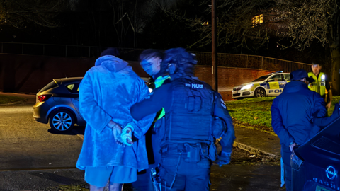 Man in handcuffs being led away by police officers after a night-time raid
