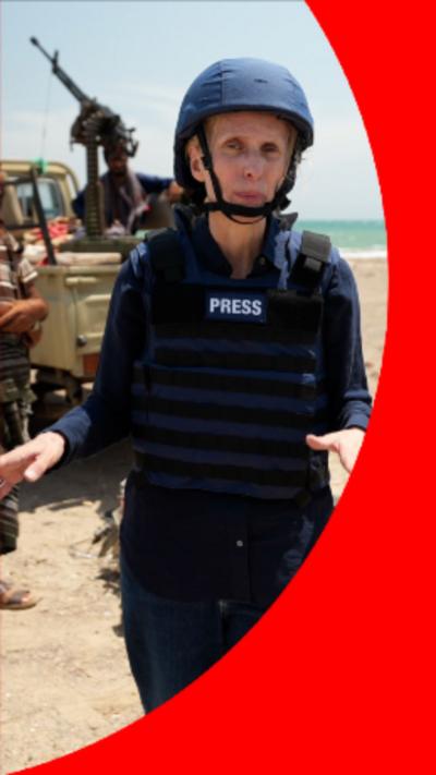 BBC Correspondent Orla Guerin is wearing a helmet with a chin-strap, and a bullet-proof vest with 'Press' written on the front. She is stood in the desert with a weapons vehicle behind her.